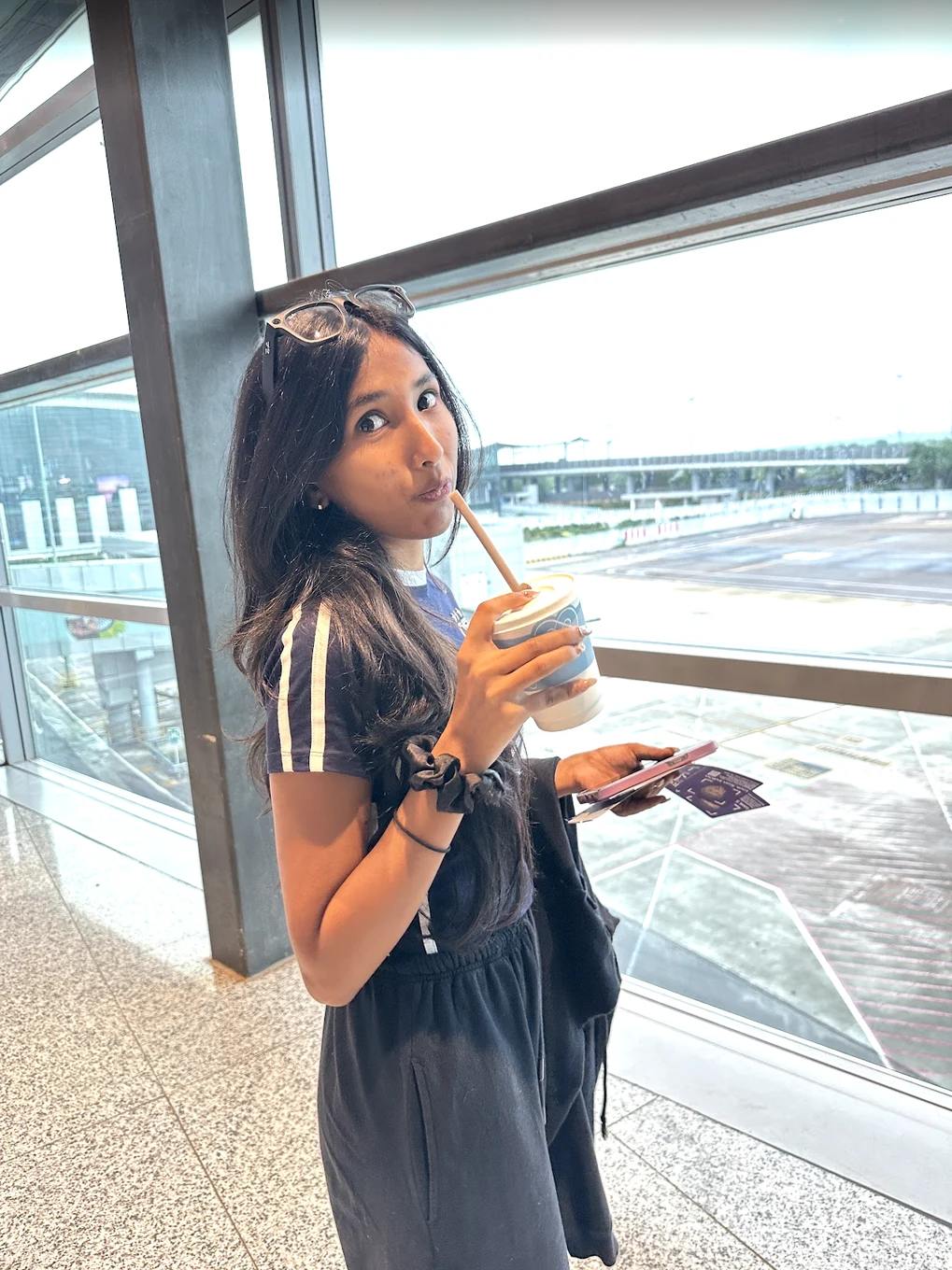 Harshitha at an airport window holding a drink.