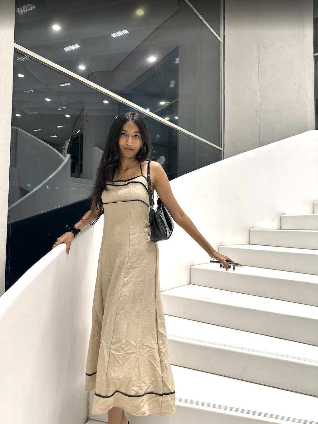 Harshitha in a cream dress with black trim, standing beside a staircase.